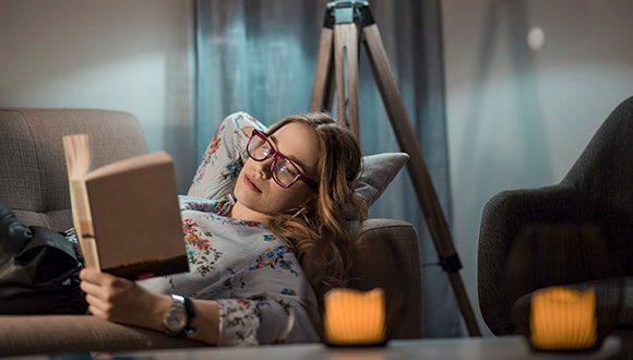 Lady laying on the lounge reading a book wearing blue light glasses