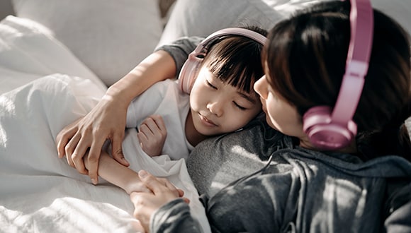 Building healthy self-esteem in kids: Mother holds young daughter listening on headphones relaxing on sofa.